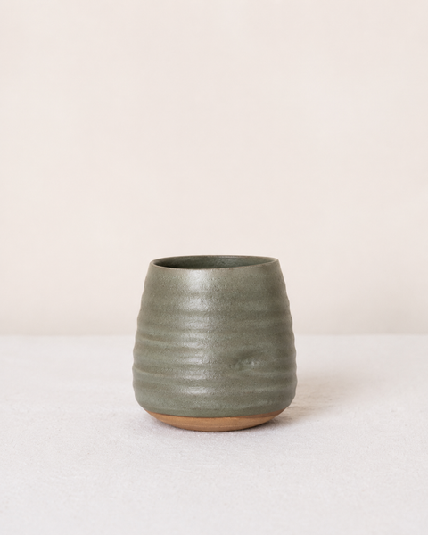 A small handmade ceramic cup with a textured, ribbed surface in a muted sage green glaze, featuring a natural unglazed clay base, set against a minimalist cream background.