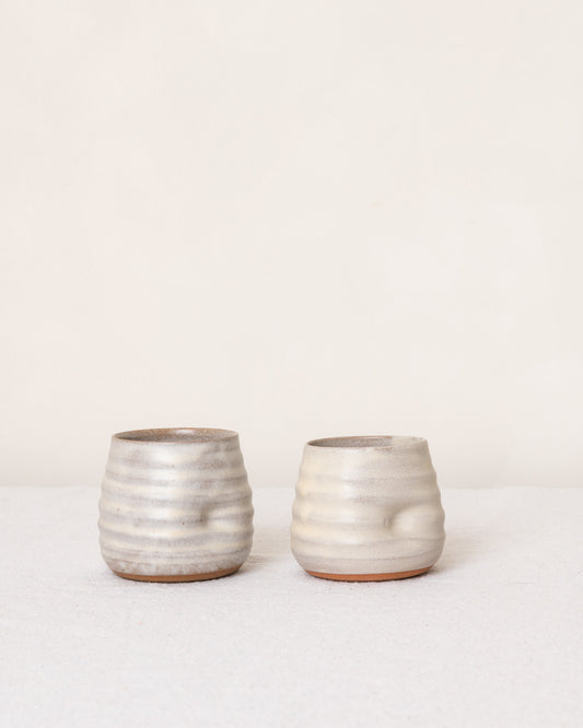 Two small ribbed ceramic cups with a textured, speckled off-white glaze and a subtle terracotta base, displayed on a light surface against a plain cream background.