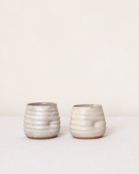 Two small ribbed ceramic cups with a textured, speckled off-white glaze and a subtle terracotta base, displayed on a light surface against a plain cream background.